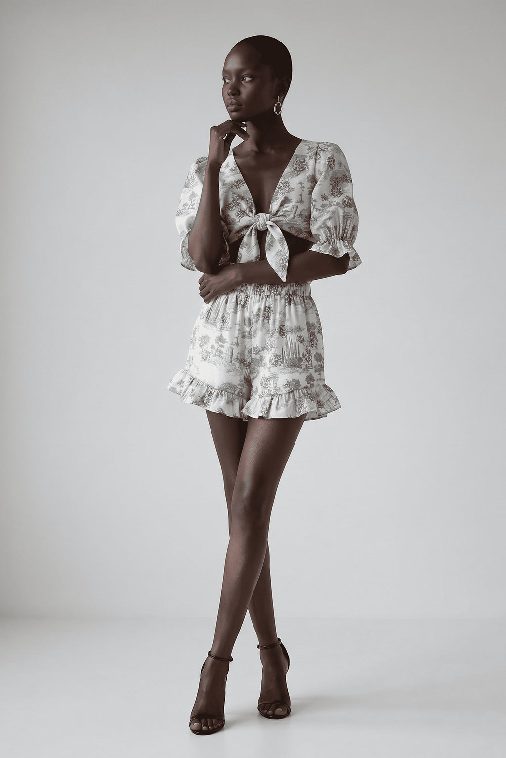 Model wearing The Saint 54 grey toile print two-piece set with tie-front puff sleeve top and ruffled mini shorts.
