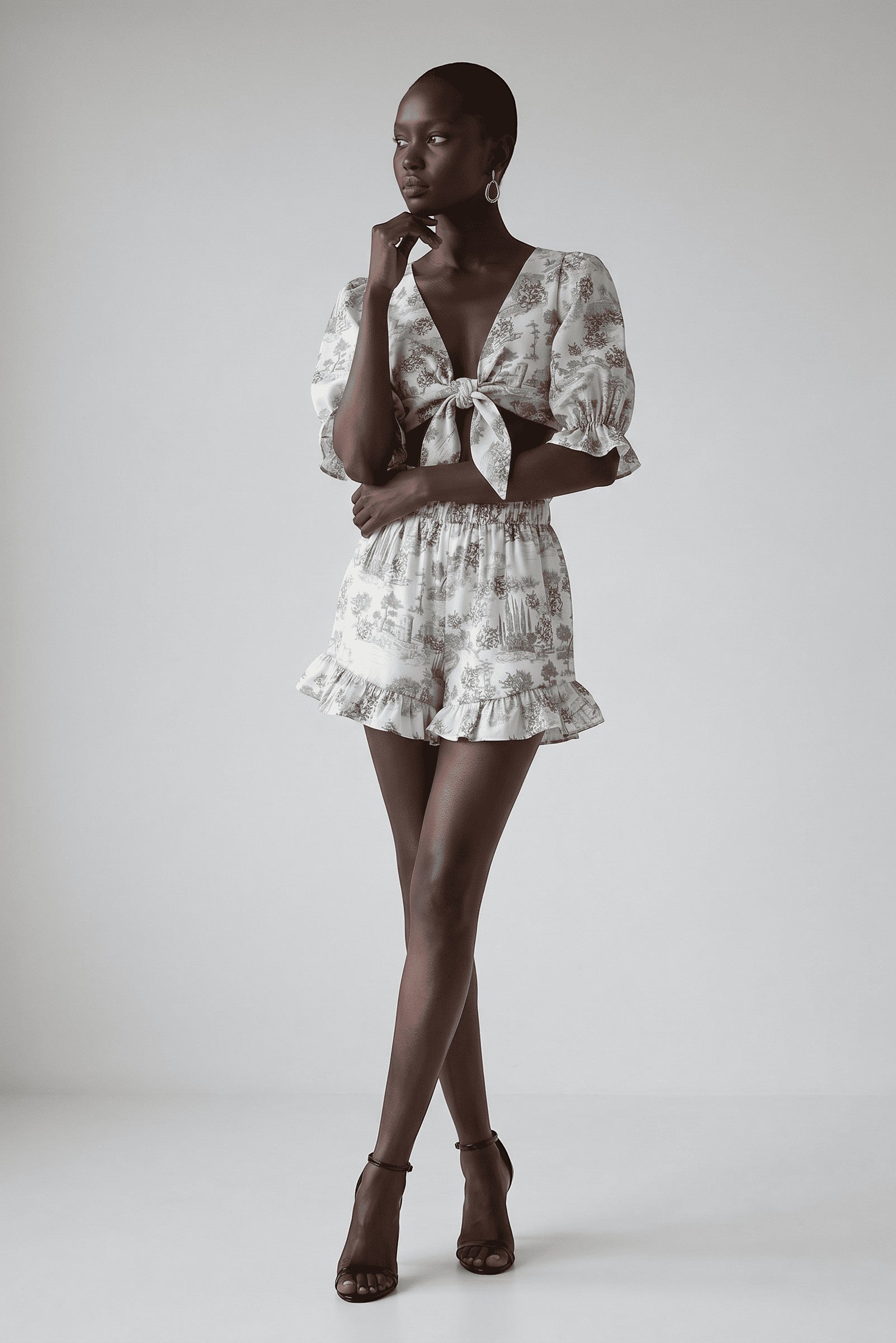 Model wearing The Saint 54 grey toile print two-piece set with tie-front puff sleeve top and ruffled mini shorts.
